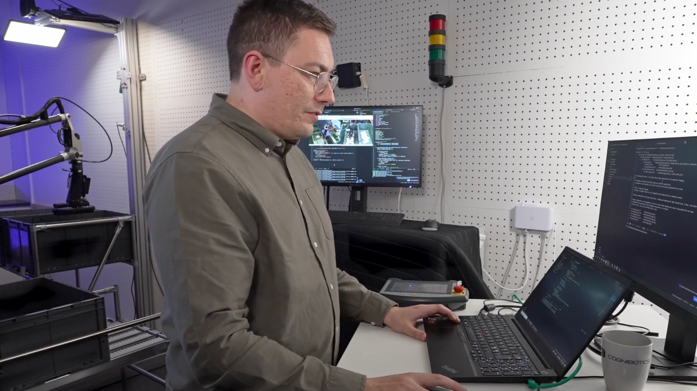 A engineer working at a desk with a laptop and monitors displaying robot code, in a tech workspace with gadgets, robot, and equipment around.