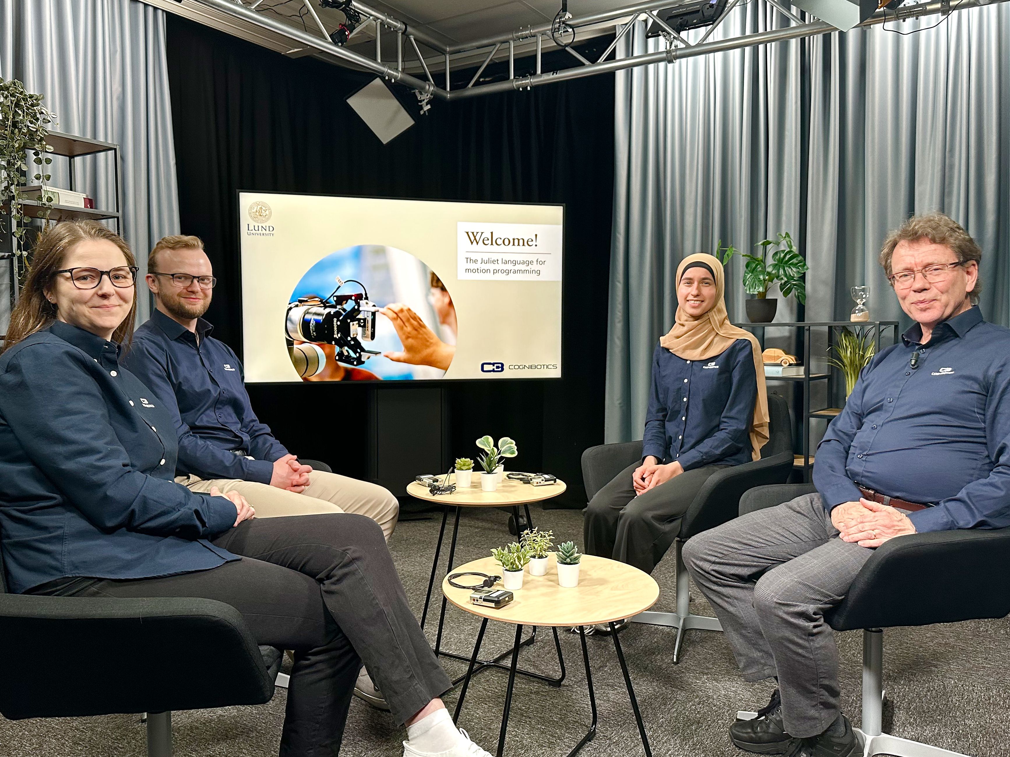 Four people seated in a studio setting, recording Juliet motion programing course, with a welcome presentation on a screen behind them, Lund University and Cognibotics
